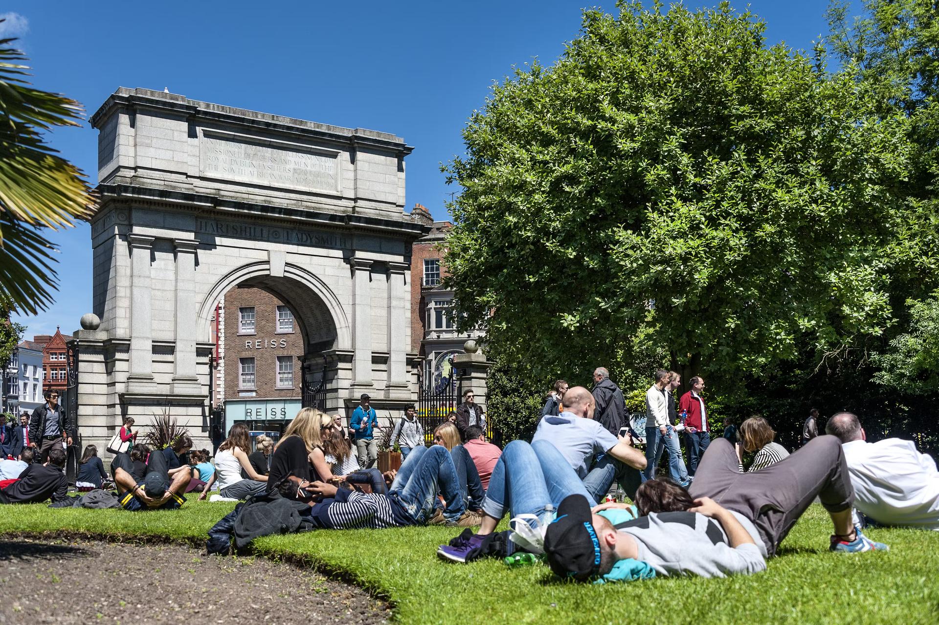 人们在都柏林的圣史蒂芬绿色的阳光明媚的日子里休息在福斯利尔拱门旁边的草地上